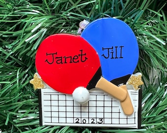 May include: A Christmas tree decoration with a ping pong theme. It features a red paddle with the name "Janet" and a blue paddle with "Jill". A white ball sits above a net, with the year "2023" displayed below.