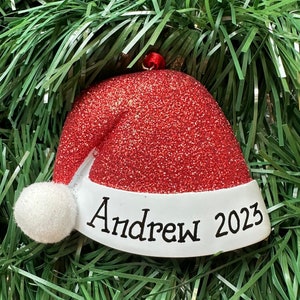 Puede incluir: Adorno navideño con forma de gorro de Papá Noel, con acabado rojo brillante y borde blanco. El nombre "Andrew" y el año "2023" están escritos en negro sobre el borde blanco. Lleva un pequeño pompón blanco.
