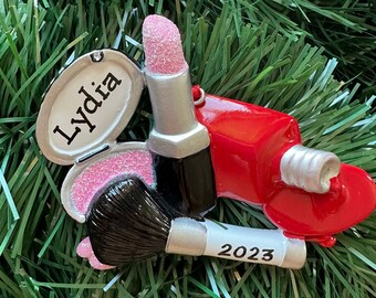 May include: A makeup-themed ornament featuring a pink lipstick, a red nail polish bottle, a makeup brush, and a compact with the name "Lydia". The brush has "2023" printed on it. The ornament is set against a green background.