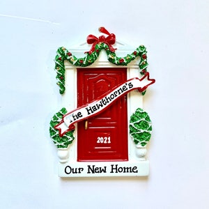 May include: Red front door Christmas ornament with a white banner that reads "The Hawthorne's" and the year "2021". The ornament also has the text "Our New Home" at the bottom.