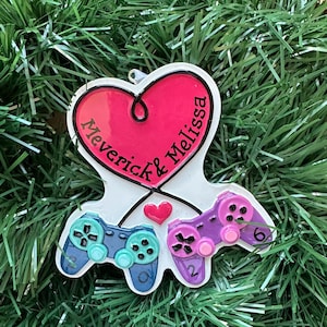 May include: A white heart-shaped ornament featuring a pink center with the text "Maverick & Melissa" in black script. Below the heart are two video game controllers, one teal and one purple, with a small pink heart between them and the numbers 0, 2, and 6.