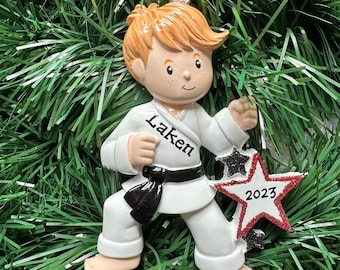 May include: A karate-themed Christmas ornament. The ornament depicts a young person in a white karate uniform with a black belt. The name "Laken" is printed on the uniform. A red and white star-shaped ornament with "2023" is attached.