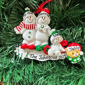 Puede incluir: Adorno navide&ntilde;o personalizado con tres mu&ntilde;ecos de nieve y un gato. Los mu&ntilde;ecos de nieve llevan sombreros y bufandas, con nombres en sus sombreros. El adorno dice "The Johnsons 2025". La figura del gato lleva un gorro de Pap&aacute; Noel y un traje verde.