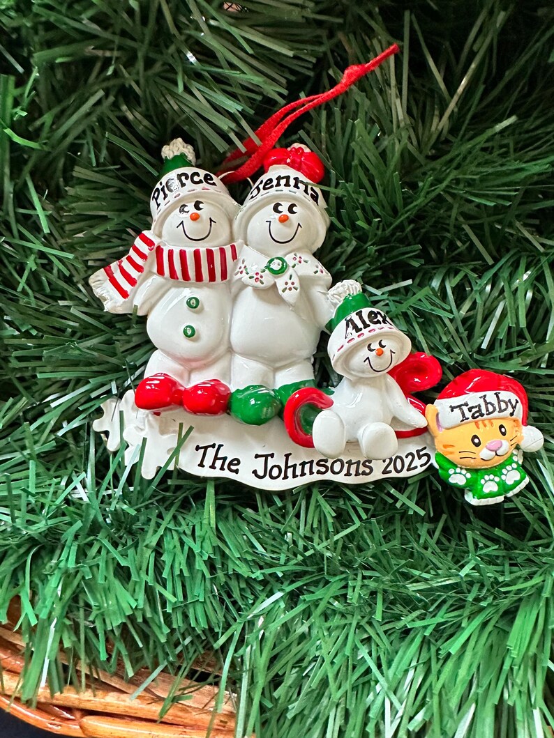 Puede incluir: Adorno navide&ntilde;o personalizado con tres mu&ntilde;ecos de nieve y un gato. Los mu&ntilde;ecos de nieve llevan sombreros y bufandas, con nombres en sus sombreros. El adorno dice "The Johnsons 2025". La figura del gato lleva un gorro de Pap&aacute; Noel y un traje verde.
