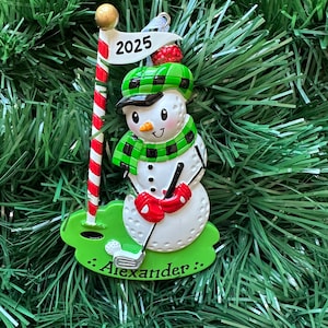 May include: A snowman Christmas ornament with a golf theme. The snowman wears a green plaid hat and scarf, red mittens, and holds a golf club. A golf flag with "2025" and the name "Alexander" is also featured. The ornament is set against a green Christmas tree.