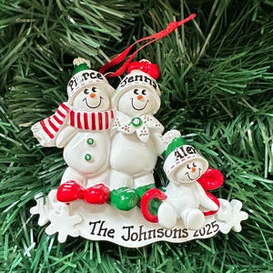 Puede incluir: Adorno navide&ntilde;o de una familia de mu&ntilde;ecos de nieve. El adorno presenta tres mu&ntilde;ecos de nieve con los nombres "Pierce", "Jenna" y "Alex" en sus sombreros. La base dice "The Johnsons 2025". Los mu&ntilde;ecos de nieve tienen detalles rojos y verdes.