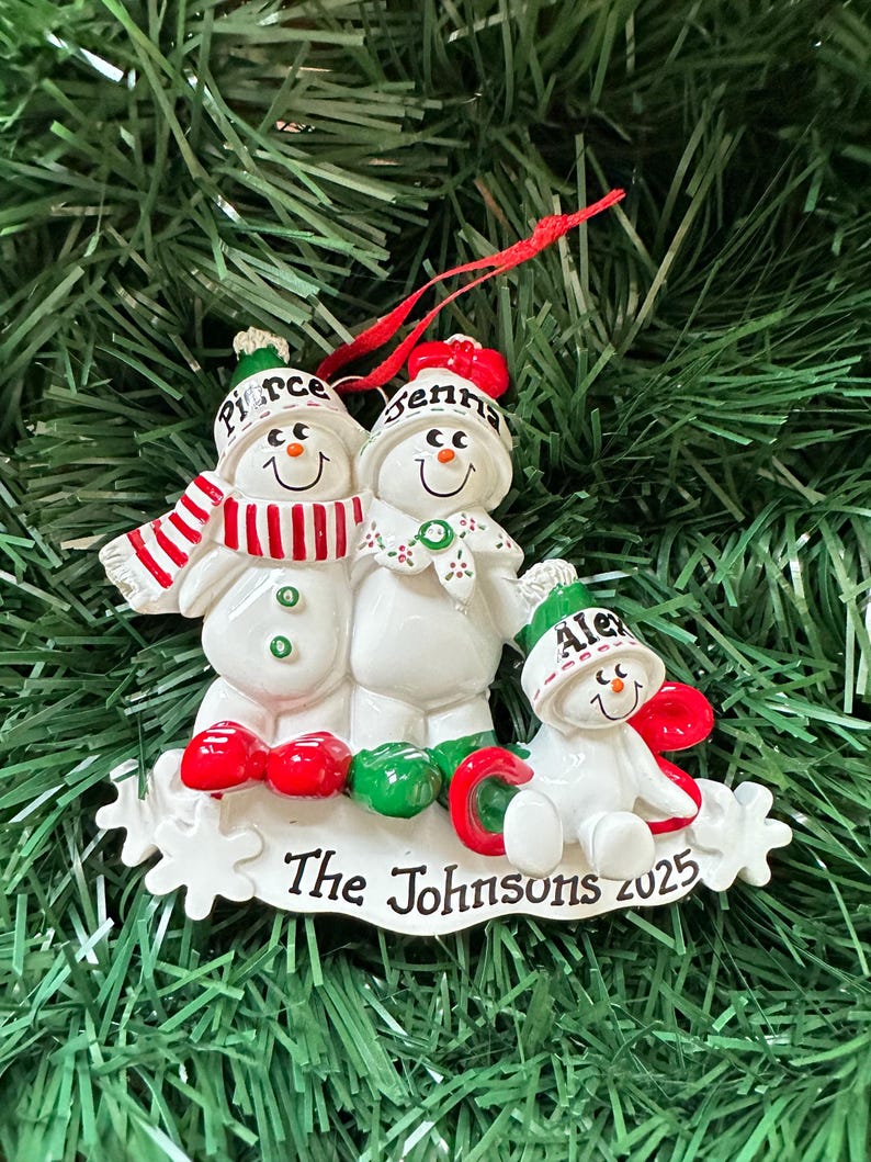 Puede incluir: Adorno navide&ntilde;o de una familia de mu&ntilde;ecos de nieve. El adorno presenta tres mu&ntilde;ecos de nieve con los nombres "Pierce", "Jenna" y "Alex" en sus sombreros. La base dice "The Johnsons 2025". Los mu&ntilde;ecos de nieve tienen detalles rojos y verdes.