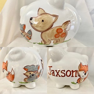 Può includere: Salvadanaio in ceramica bianca con tema di animali del bosco. Il salvadanaio presenta una volpe, un procione e un cervo. Il nome Jaxson è scritto sul lato del salvadanaio in arancione.