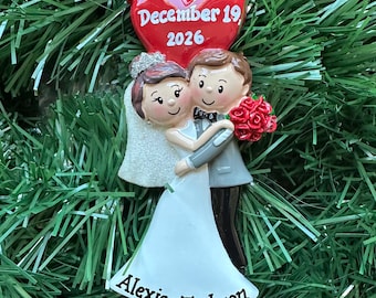 May include: A wedding ornament featuring a bride in a white dress and a groom in a grey suit, embracing. Above them is a red heart with the text December 19, 2026. The base of the ornament displays the names Alexis and Jackson with a small red heart.