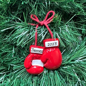 May include: Red boxing glove Christmas ornament with the name "James" on one glove and the year "2023" on the other. The ornament hangs from a red string and is set against a backdrop of green Christmas tree branches.