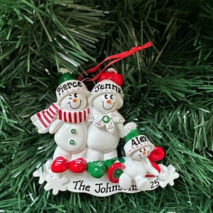 Puede incluir: Adorno navide&ntilde;o de una familia de mu&ntilde;ecos de nieve. El adorno presenta tres mu&ntilde;ecos de nieve con los nombres "Pierce", "Jenna" y "Alex" en sus sombreros. La base dice "The Johnsons 25". Los mu&ntilde;ecos de nieve son blancos con detalles rojos y verdes.