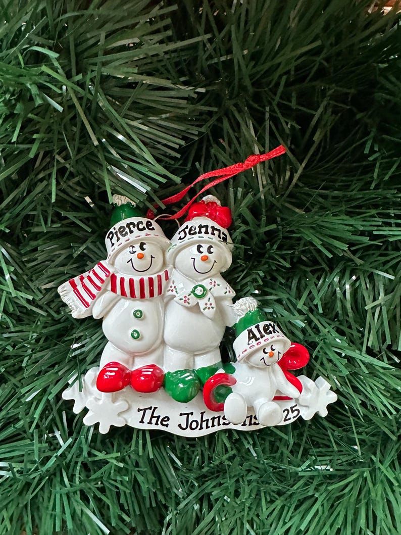 Puede incluir: Adorno navide&ntilde;o de una familia de mu&ntilde;ecos de nieve. El adorno presenta tres mu&ntilde;ecos de nieve con los nombres "Pierce", "Jenna" y "Alex" en sus sombreros. La base dice "The Johnsons 25". Los mu&ntilde;ecos de nieve son blancos con detalles rojos y verdes.