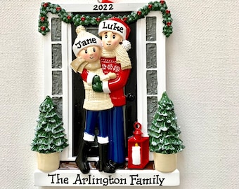 May include: A Christmas ornament featuring two figures, Jane and Luke, standing in front of a black door. The ornament displays the year 2022 at the top and the text The Arlington Family at the bottom, framed by green garlands and two small pine trees.