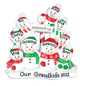 May include: A festive Christmas ornament depicting snowmen with names such as Alex and Mia. The snowmen are adorned with red and green scarves and hats. The ornament includes the text "North Pole" and "Our Grandkids 2022".