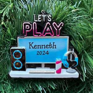 May include: A Christmas ornament with a gaming theme. The ornament features a computer monitor with the name "Kenneth" and the year "2024". It includes a speaker, headphones, a keyboard, and a drink. The words "LET'S PLAY" are at the top.