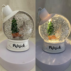 Könnte beinhalten: Eine weiße und transparente Schneekugel mit einem Weihnachtsbaum und einem Zwerg im Inneren. Auf dem Sockel der Kugel steht in schwarzer Schrift "Patrick". Die Kugel wird von innen beleuchtet und erzeugt ein warmes Leuchten.