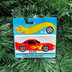 May include: A red car ornament with yellow flames and black wheels. The ornament has the text "2025 Jackson" on a white background, set against a blue backdrop with a yellow flame and wheel design. The ornament is hanging on a green, artificial Christmas tree.