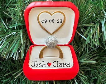 Op de afbeelding: Rode en witte kerstversiering in de vorm van een open ringdoos met een glinsterende ring erin. De doos heeft een gouden hart met de datum 09-08-23 en de namen Josh en Clara met een hart ertussen.