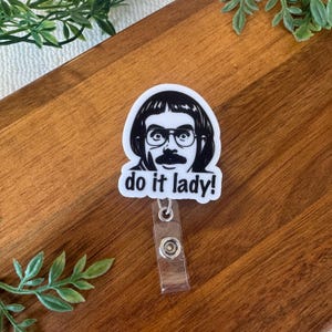 May include: A white badge reel with a black and white graphic of a man with glasses and a mustache. The text "do it lady!" is below the graphic. The badge reel has a clear plastic strap and a metal clip.