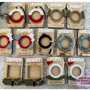 May include: A collection of wooden Father's Day photo frames with various designs and text. The frames feature words like "POPS", "UNCLE", "GRANDPA", and "DADDY". Each frame has a circular or rectangular opening for a photo, with decorative elements.