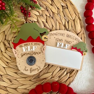 May include: Wooden Christmas elf surveillance sign with a green elf hat, red trim, and a camera lens. The sign reads "This house is under Elf Surveillance" and "The Berry's". A second sign reads "Important Message from your Elf".