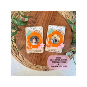 May include: Two wooden Mother's Day photo frames with orange accents. One says "Mommy" and the other "Grandma." Each frame has an orange circle, a photo, and the text "Happy Mother's Day! From your little cutie."
