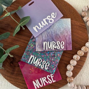May include: Four square acrylic badge reels featuring the word nurse in white script or block lettering. The top is solid lavender, the second has a purple and blue marbled finish, the third has iridescent glitter, and the bottom is solid magenta.