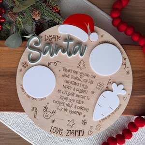 May include: A round wooden Christmas Eve plate with a Santa hat, two white circles, and a carrot shape. The plate has the words "Dear Santa" and festive text, including "Love, Zianni". Red felt ball garland is on the right.