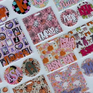 May include: A collection of square and circular acrylic badge reels featuring various patterns including pumpkins, ghosts, bows, and checkered prints. Several pieces display raised lettering such as nurse, BSN, RN, PEDS, and various initials.