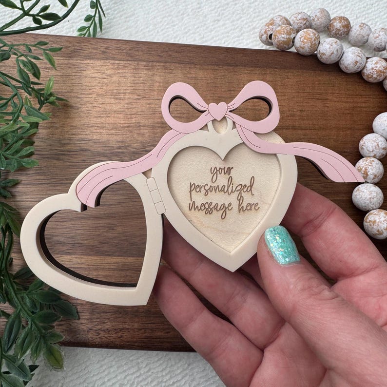 May include: A heart-shaped wooden frame with a hinged opening, a light pink bow, and the text "your personalized message here." The frame is open, revealing a space for a photo or message. The frame is held by a person.