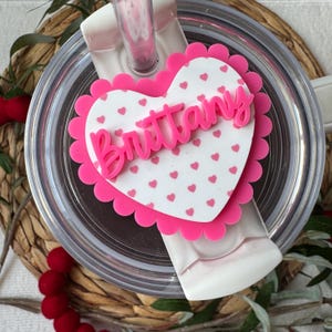 Op de afbeelding: Een roze hartvormig label met de naam "Brittany" in cursief. Het label heeft een geschulpte rand en een witte achtergrond met kleine roze hartjes. Het label is bevestigd aan een doorzichtig tumblerdeksel.