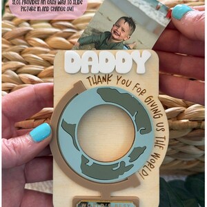 May include: Wooden photo holder with the word "DADDY" in white letters. The holder features a globe design and the text "THANK YOU FOR GIVING US THE WORLD!" and "WORLD'S BEST DAD". A 2x3 inch photo is inserted into the slot.