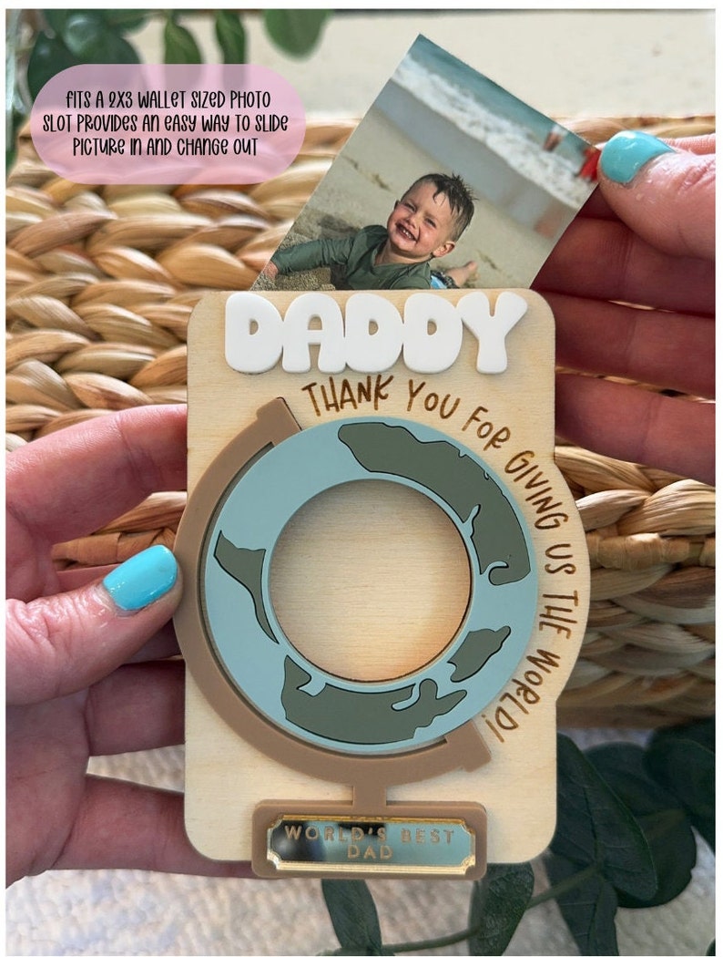 May include: Wooden photo holder with the word "DADDY" in white letters. The holder features a globe design and the text "THANK YOU FOR GIVING US THE WORLD!" and "WORLD'S BEST DAD". A 2x3 inch photo is inserted into the slot.