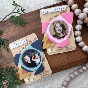 May include: Two wooden photo frames with rocket ship cutouts. One frame says "Nana" and the other says "Grammy". Each frame has a photo of a child. The frames have the text "We love you to the moon" and "Love, Bentley & Braxton" and "Love, Teagen".