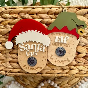 以下が含まれることがあります： 2つの木製クリスマスオーナメント。1つは「サンタカム」の文字が入ったサンタの頭の形、もう1つは「エルフ監視」の文字が入ったエルフの頭の形です。どちらにもカメラレンズと家族の名前が入っています。赤、白、緑、茶色の色。