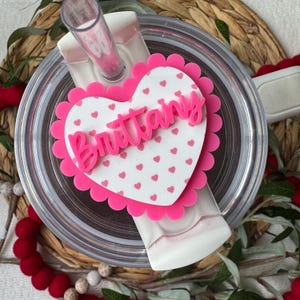 Op de afbeelding: Een doorzichtige beker met een roze hartvormige topper. De topper heeft een geschulpte rand en de naam "Brittany" in roze script. Het hart is wit met kleine roze hartjes verspreid. Een roze rietje is zichtbaar.