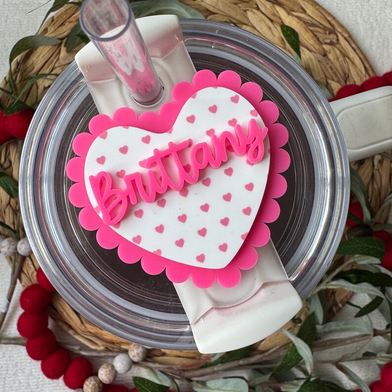 Op de afbeelding: Een doorzichtige beker met een roze hartvormige topper. De topper heeft een geschulpte rand en de naam "Brittany" in roze script. Het hart is wit met kleine roze hartjes verspreid. Een roze rietje is zichtbaar.