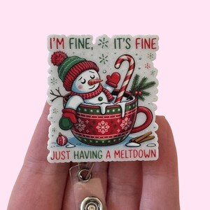 May include: A festive badge reel featuring a snowman in a red and green Christmas mug with a candy cane. The text reads "I'm fine, it's fine, just having a meltdown". The reel has a clear plastic strap and a metal button.