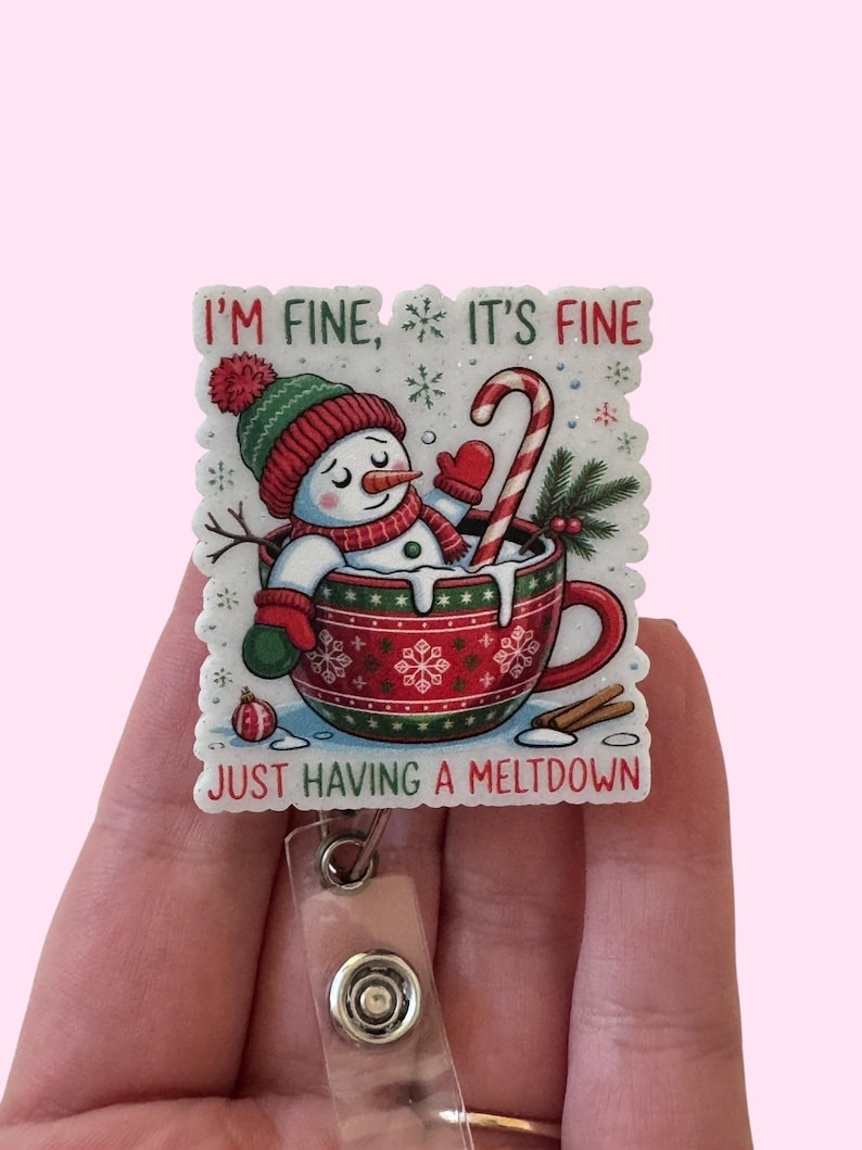 May include: A festive badge reel featuring a snowman in a red and green Christmas mug with a candy cane. The text reads "I'm fine, it's fine, just having a meltdown". The reel has a clear plastic strap and a metal button.