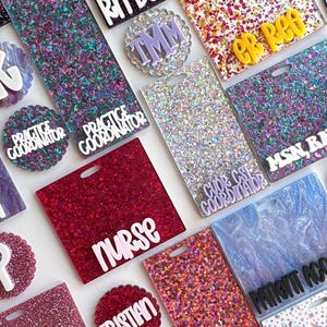 May include: A collection of rectangular and circular acrylic badge reels featuring various glitter finishes in purple, pink, silver, and multi-colored patterns. Each piece displays white or yellow raised lettering with medical titles like RN BSN, NURSE, CCMA, and X-RAY.
