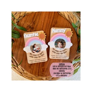 May include: Two wooden Mother's Day cards with rainbow and sun designs. One card says "Auntie" and the other says "Mommy". Each card has a photo and a message: "You make us happy when skies are grey."