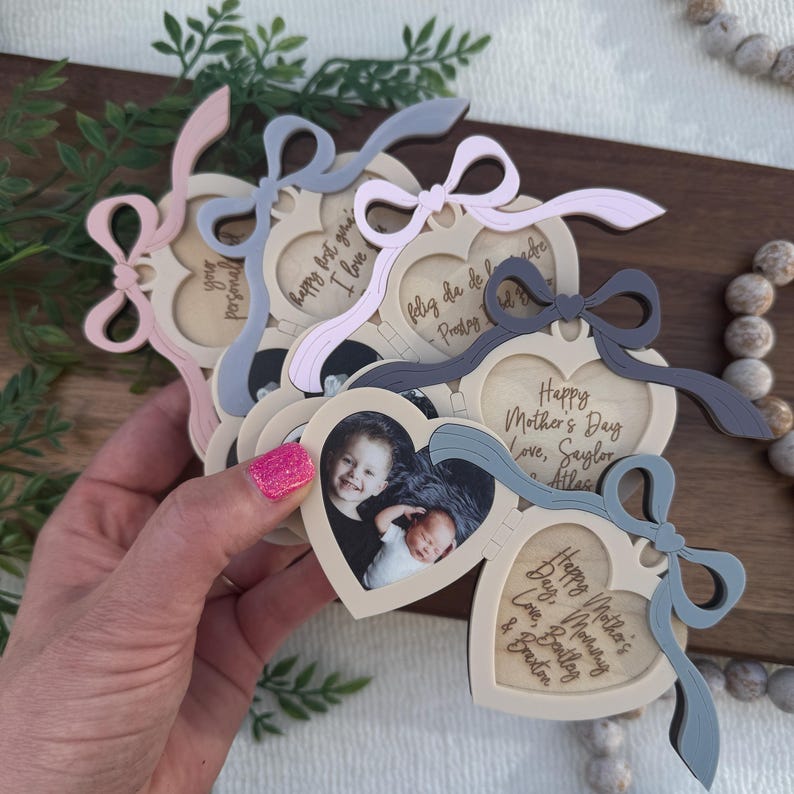 May include: A collection of heart-shaped photo frames with attached bows in various pastel colors. Each frame holds a small photograph and includes engraved text such as "Happy Mother's Day" and personalized names. The frames are held in a hand.