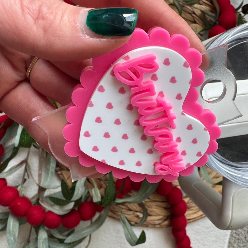 Op de afbeelding: Een roze en witte hartvormige badge met de naam "Brittany" in roze script. Het hart heeft een geschulpte rand en een patroon van kleine rode hartjes op een witte achtergrond. Een doorzichtige plastic clip is bevestigd.