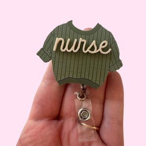 Puede incluir: Un portatarjetas en forma de suéter verde con la palabra "nurse" en escritura color crema. El portatarjetas tiene una correa de plástico transparente y un clip de metal.