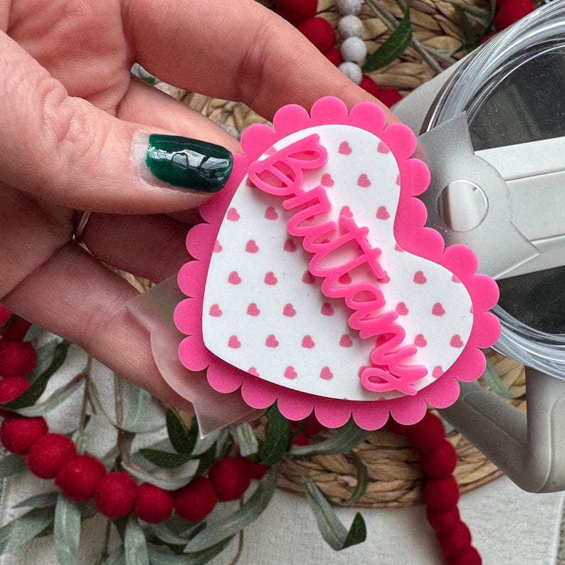 Op de afbeelding: Een roze hartvormig accessoire met een geschulpte rand en de naam "Brittany" in cursief. Het hart heeft een witte achtergrond met kleine roze hartjes. Het accessoire wordt vastgehouden door een hand met een groene nagel.