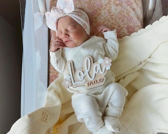 May include: A newborn baby wearing a white onesie, matching hat, and booties, lying on a cream-colored blanket. A wooden name sign with the name "Lola mae" is placed on the onesie. The baby is in a clear bassinet with a floral patterned background.