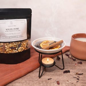 Peut inclure: Un sachet noir avec l'inscription "Simmer Pot Soleil Dore" à côté d'un bol blanc contenant des tranches d'orange séchées et une cannelle, sur un support en métal noir avec une bougie chauffe-plat allumée. Une bougie en terre cuite est à droite.