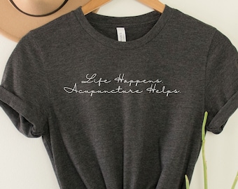 May include: A dark grey t-shirt with the text "Life Happens, Acupuncture Helps" in white lettering.