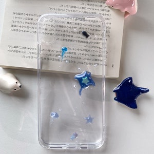 以下が含まれることがあります： 青と緑の海の生き物、マンタやクラゲで飾られた透明な携帯電話ケース。 ケースは水っぽく泡立った効果があり、小さく透明な装飾が施されています。 水中のシーンを連想させるデザインです。