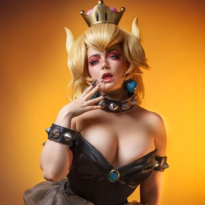 May include: A woman dressed as Princess Peach from the Super Mario video game series. She is wearing a black and gold dress with a crown and a spiked collar. She has blonde hair and blue eyes. The background is a solid orange color.